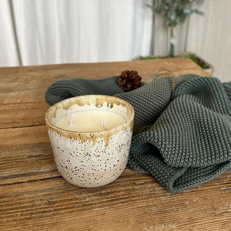 Duftkerze handgemacht im Keramikbecher auf einem Holztisch mit grünem Stoff und einer Tannenzapfen-Dekoration.